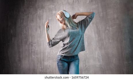 Joven mujer rubia positiva en un suéter gris de gran tamaño bailando música en auriculares, ventilador de música