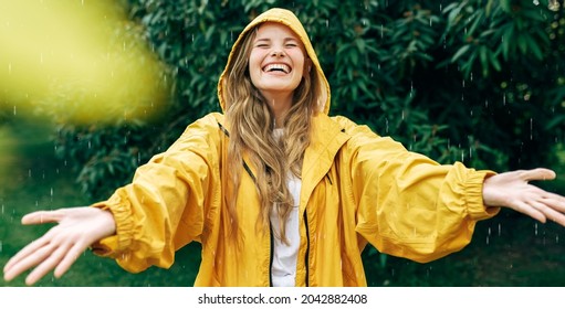 Positive young blonde woman smiling wearing yellow raincoat during the rain in the park. Cheerful female enjoying the rain outdoors. A beautiful woman catching the raindrops with arms wide open. - Powered by Shutterstock