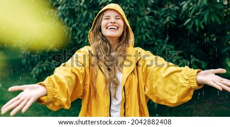 Positive young blonde woman smiling wearing yellow raincoat during the rain in the park. Cheerful female enjoying the rain outdoors. A beautiful woman catching the raindrops with arms wide open.