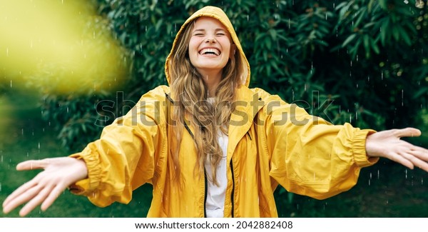 Positive young blonde woman smiling wearing yellow\
raincoat during the rain in the park. Cheerful female enjoying the\
rain outdoors. A beautiful woman catching the raindrops with arms\
wide open.