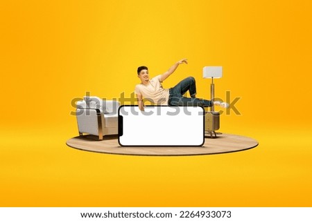 Positive young boy lying on huge 3D model of mobile phone with empty screen for text, ad over bright yellow background with home interior. Online shopping, delivery. Mockup for ad, design, logo.