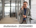 Positive young businessman in glasses discussing over mobile phone while standing in office corridor