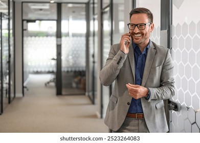 Positive young businessman in glasses discussing over mobile phone while standing in office corridor - Powered by Shutterstock