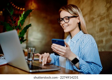 Positive young businesswoman in casual clothes and eyeglasses sitting in cafe with cellphone while working on remote project using laptop - Shutterstock ID 2274914231