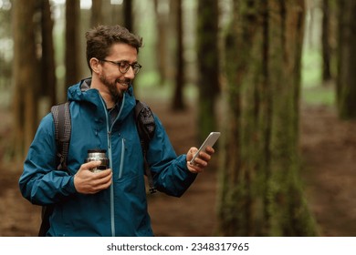 Positive young caucasian couple in jackets with cup of coffee, walk in forest together, enjoy trip, use phone map, outdoor. Hiking, lifestyle, adventure and tourism alone with device