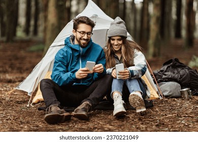 Positive young caucasian family in jackets sitting near tent, chatting on smartphone, enjoy adventure, spare time at weekend, travel blog app in cold forest in autumn. Vacation together, lifestyle - Powered by Shutterstock