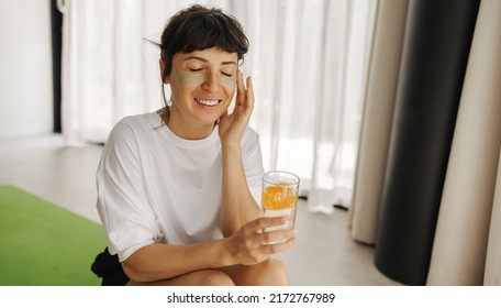 Positive Young Caucasian Girl With Patches And Water Moisturizes Herself Inside And Outside Sitting At Home. Brunette Woman With Closed Eyes Wears Casual Summer Clothes. Wellness And Self Care Concept