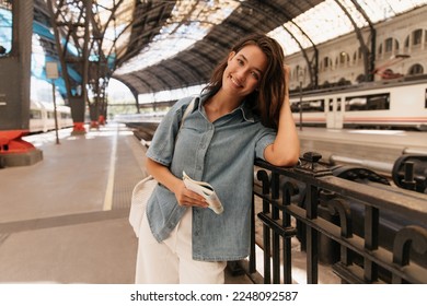 Positive young caucasian girl posing looking at camera spends time at railway station alone. Brunette wears casual clothes and bag. Concept of tourism.