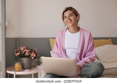 Positive young caucasian girl with wireless headphones and laptop sitting on bed indoor. Smiling woman wears casual clothes at home. Concept gadgets, technologies. - Powered by Shutterstock