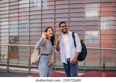 Positive Young Caucasian Interracial Young Guy With Girl Waving Hands Standing On Street. Students Wearing Casual Clothes Take Break Outside College. 