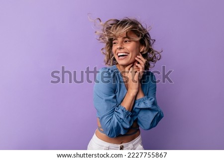 Positive young caucasian lady with tousled hair smiles broadly teeth on purple background. Blonde looks away, wears blue blouse. Emotion concept