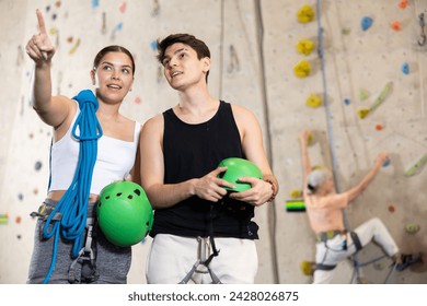 Positive young couple standing with blue safety belts and helmet before practicing rock-climbing on climbing wall in adventure park - Powered by Shutterstock