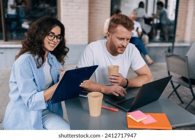 Positive young coworkers smilingб sitting at table with laptop smartphone cup of coffee notebook while male looking at screen of laptop and female on clipboard while working on remote project in cafe - Powered by Shutterstock