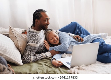 Positive young diverse couple looking at laptop while relaxing on bed - Powered by Shutterstock