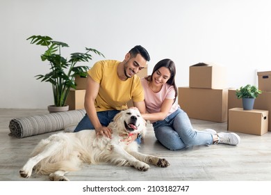 Positive Young Diverse Couple Stroking Their Dog In New Home On Moving Day, Full Length Portrait. Millennial Multiracial Family Playing And Cuddling With Cute Pet Indoors. Home Relocation Concept