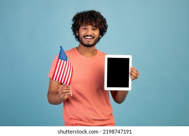 Positive Young Eastern Man In Casual Student Showing Flag Of The US And Modern Digital Tablet With Black Blank Screen, Recommending Educational Mobile App On Blue Studio Background, Mockup