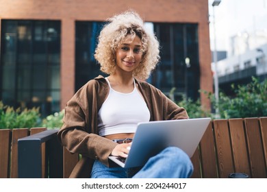 Positive young ethnic female freelancer in casual clothes sitting on wooden bench and browsing laptop while working on remote project