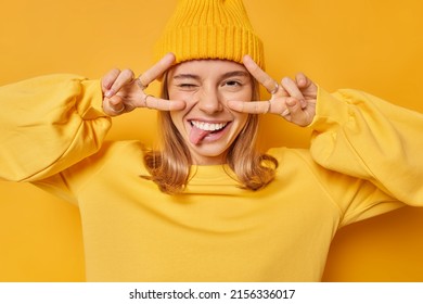 Positive young European woman makes peace gesture over eyes sticks out tongue demonstrates victory sign wears hat and casual jumper isolated over vivid yellow background. Body language concept - Shutterstock ID 2156336017