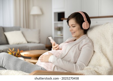 Positive young expectant mom in pink wireless headphones chatting online, texting on smartphone, listening to music, virtual playlist for stress relief and relaxation. Pregnant woman enjoying leisure - Powered by Shutterstock