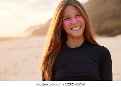 Positive young experienced surfer smiles broadly at camera, squits face with pleasure, protects face zinc dioxide, wears diving suit, ready for surfing across ocean, poses for womens magazine