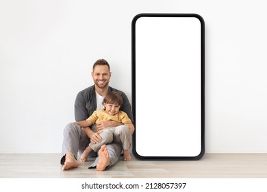 Positive Young Father Embracing His Little Son, Sitting Together On Floor Near Big Smartphone With Blank Screen Against White Studio Wall, Mockup For Online Advertisement, Mobile App Or Website Design