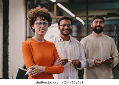 Positive young female architect in curly hair and eyeglasses with multiracial coworkers in casual clothes smiling and looking at camera while standing in modern workspace - Powered by Shutterstock