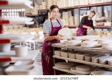 Positive young female artisan focused on stacking newly crafted ceramic plates on racks for even drying in spacious pottery workshop - Powered by Shutterstock