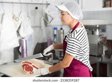 Positive young female butcher shop worker wearing red apron and mesh cap with visor, cutting slab of beef with large knife in processing room, preparing meat products for sale - Powered by Shutterstock