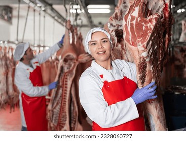 Positive young female butcher working in chilling room of meat processing factory, arranging raw beef carcasses hanging on hooks for storage - Powered by Shutterstock