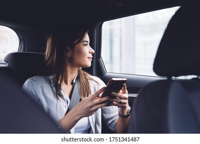 Positive Young Female In Casual Clothes Using Mobile Phone While Sitting In Comfortable Car Backseat And Looking At Window Dreamily