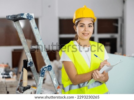 Positive young female civil engineer wearing yellow vest and safety hard hat taking notes while checking indoor construction site, satisfied with progress of work..