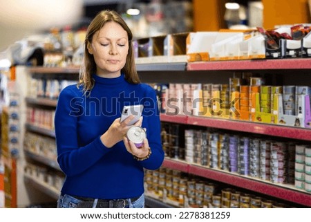 Positive young female customer scanning barcode on tin can of canned food for cats with smartphone while shopping in pet store, paying for item using mobile app..
