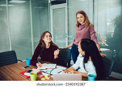 Positive young female employee sitting with unrecognizable coworker and looking at each other at table with papers smartphones cups laptop and discussing details of project while smiling woman - Shutterstock ID 2340192387