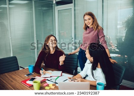 Positive young female employee sitting with unrecognizable coworker and looking at each other at table with papers smartphones cups laptop and discussing details of project while smiling woman
