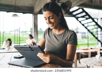 Positive young female freelancer in casual t shirt smiling and browsing tablet while using stylus during remote work from cafe - Powered by Shutterstock