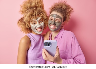 Positive Young Female Friends With Curly Hair Applies Beauty Mask For Skin Treatment Use Mobile Phone Dressed In Casual Clothes Take Selfie Isolated Over Pink Background. Facial Care Concept