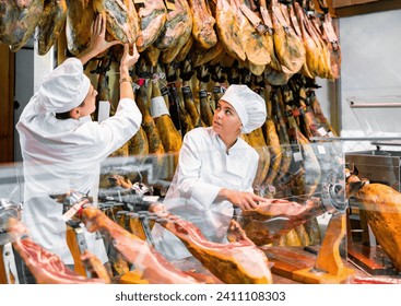 Positive young female and male vendors working at counter in butcher shop, arranging dry-cured spanish jamon for sale