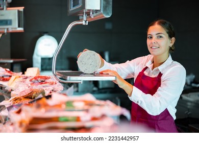 Positive young female seller of local grocery store standing behind counter, weighing traditional spanish mortadella on scales - Powered by Shutterstock