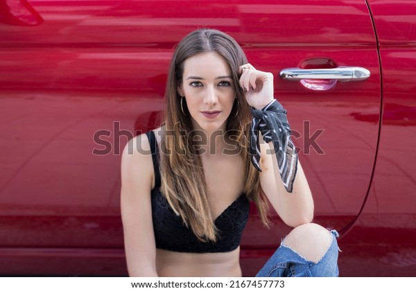 Positive young\
female in trendy outfit with long hair looking at camera while\
standing near red car on city\
street