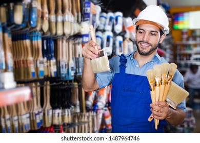 Positive Young Foreman In Blue Overalls Choosing Brushes In Paint Store