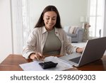 Positive young freelance accountant woman typing on calculator at home, calculating budget, income, savings, paying bills, taxes on online financial app on laptop, enjoying financial management