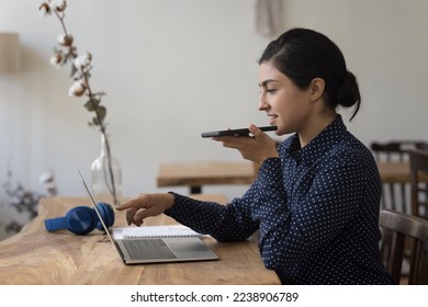 Positive young freelance Indian business woman using app on gadgets for online communication, recording voice message on smartphone, pointing at laptop computer monitor, working at desk in home office - Powered by Shutterstock