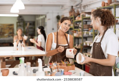 Positive young girl and guy in apron painting on handmade ceramic bowl with brush in pottery workshop - Powered by Shutterstock