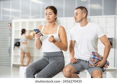 Positive young girl in gym attire holding smartphone while sitting with male friend in bouldering club locker room, taking break during climbing session.. - Powered by Shutterstock