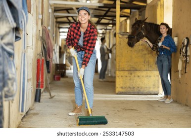 Positive young girl horse farm worker cleaning floor at private stable. Adult woman holds horse by reins in background - Powered by Shutterstock