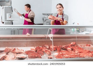 Positive young girl, professional butcher shop saleswoman, arranging meat products in refrigerated display case, laying out fresh raw beef ..