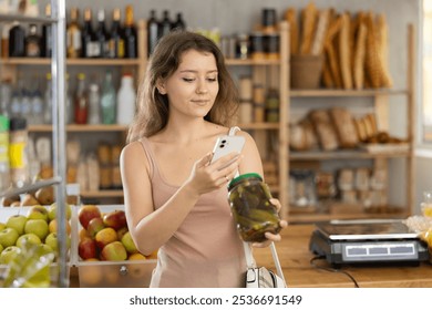 Positive young girl is scanning QR code on jar of pickled cucumber in grocery store - Powered by Shutterstock