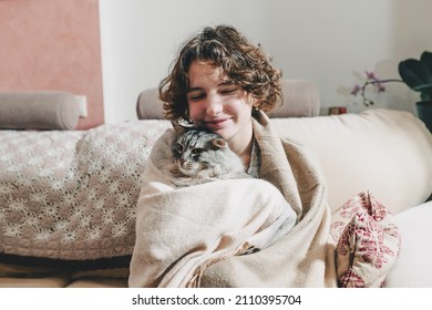 positive young girl, smiling, wrapped in warm blanket, sits on comfortable sofa, hugging purring gray fluffy cat. hugging each other, girl and domestic cat are warming themselves in cold house