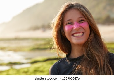 Positive Young Glad European Woman With Toothy Smile, Has Protective Zinc Mask On Face Which Blocks Sun Rays, Wears Diving Suit For Surfing, Poses Outdoor Against Blurred Coastline Background.