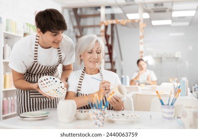 Positive young guy assisting enthusiastic senior woman painting ceramic mug in pottery class, showing plate with paint samples. Intergenerational communication and creative hobby concept - Powered by Shutterstock